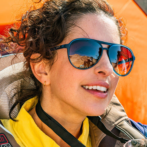 A woman wearing navy blue aviator sunglasses with dark amber lenses smiles softly looking off into the distance.